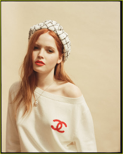 Portrait of a red-haired woman wearing a white sweater and textured headband, posing against a neutral background in soft lighting.