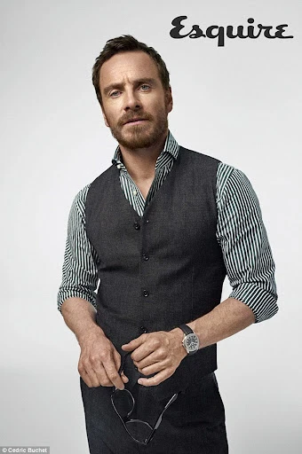 Portrait of a handsome adult man in a vest and striped shirt, holding glasses, against a neutral background with an Esquire logo.