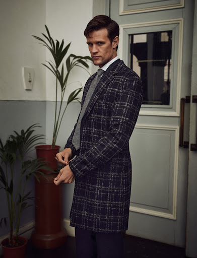 Adult male in a patterned coat stands in a neutral indoor location, looking slightly down. He wears a white shirt and grey tie.
