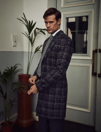 Adult male in a patterned coat stands in a neutral indoor location, looking slightly down. He wears a white shirt and grey tie.