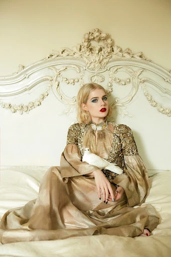 Blonde woman in an ornate gown sits on a bed holding a white dove, posing in front of a decorative cream-colored headboard in an indoor setting.