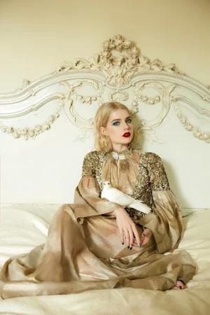 Blonde woman in an ornate gown sits on a bed holding a white dove, posing in front of a decorative cream-colored headboard in an indoor setting.