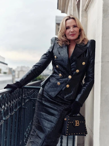 Elegant adult woman in a black suit with a handbag, standing on a balcony with city buildings visible in the background on a cloudy day.