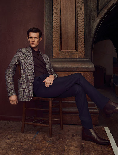 Man in a brown plaid jacket and blue trousers sitting on a wooden chair in an indoor setting with warm lighting and a serious expression.