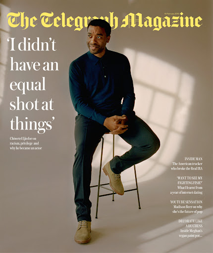 Chiwetel Ejiofor on a magazine cover, sitting on a stool in a studio. He is wearing a blue shirt and tan shoes.