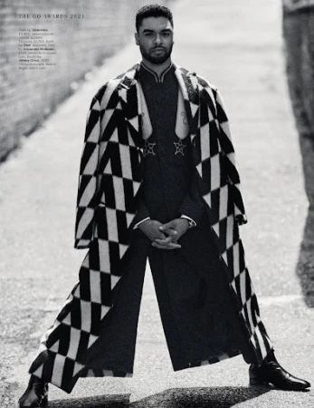 Man in black and white patterned coat stands in alleyway with confident pose, showcasing fashion in an urban outdoor setting.