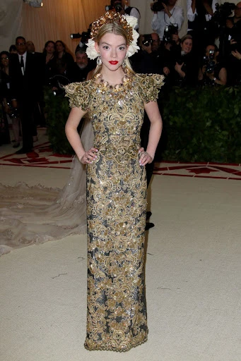 Woman in gold and black gown and flower headpiece attends event. Close-up view, indoor environment, event lighting, and large crowd visible.