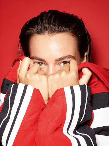 Close-up portrait of a person with slicked-back dark hair holding a red and white jacket up to cover their face.