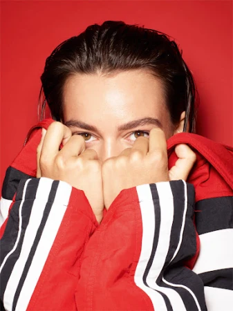Close-up portrait of a person with slicked-back dark hair holding a red and white jacket up to cover their face.
