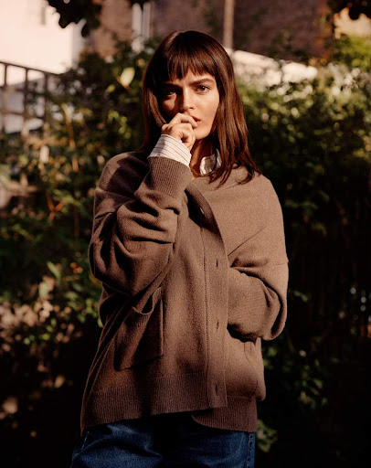 Woman in brown cardigan poses in a lush outdoor setting with sunlight, wearing denim jeans and a collared shirt for a natural, relaxed look.