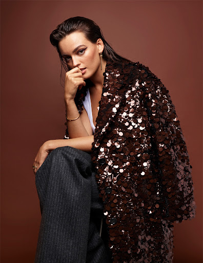 Portrait of a brunette adult woman wearing a sequined jacket and pinstripe trousers against a warm brown studio backdrop.