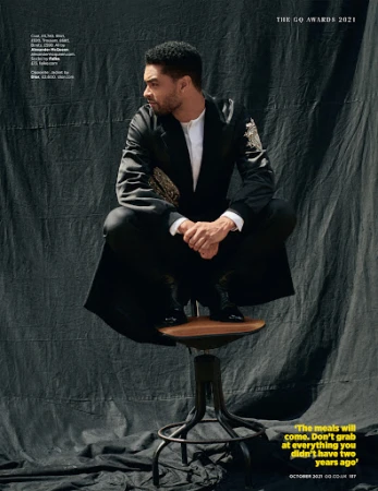 Regé-Jean Page crouched on a stool against a dark textured backdrop, wearing a satin jacket and trousers