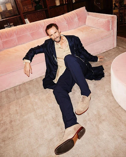 Indoor studio shot of a male model lounging on a pink sofa, wearing a navy jacket, beige shirt, blue trousers, and suede shoes.