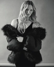 Black and white portrait of a woman with long blonde hair wearing a dark furry jacket and dark top against a plain grey studio backdrop.