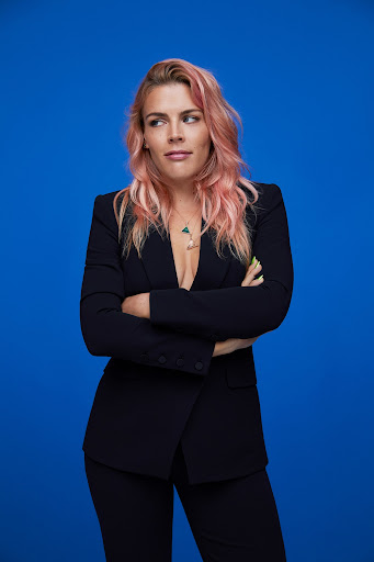 Blonde woman with pink hair in a black suit stands confidently against a bright blue background with her arms crossed. Studio shot.