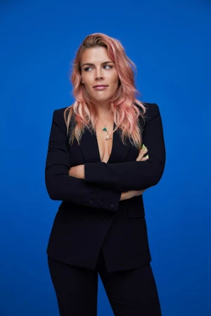 Blonde woman with pink hair in a black suit stands confidently against a bright blue background with her arms crossed. Studio shot.