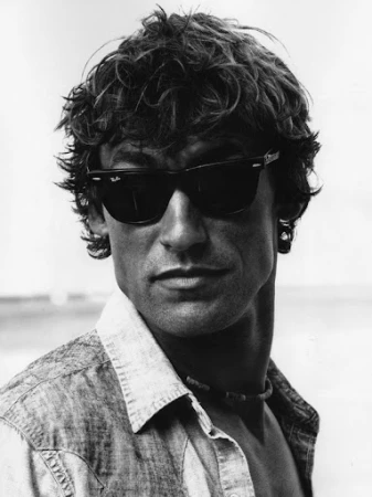 Portrait of a man with curly hair and dark sunglasses wearing an unbuttoned patterned shirt against a plain background.