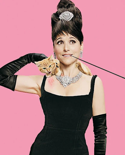 Adult female in black dress and gloves with updo hairstyle, decorative jewelry, holding long cigarette holder, and kitten on shoulder.