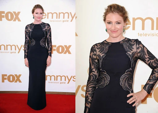 A woman in a long black dress with lace sleeves poses on a red carpet in front of Emmy Awards signage. She smiles subtly at the camera.