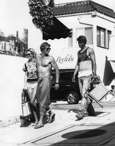 Three people walk on a sidewalk near a building. One man carries a folding chair. The image is in black and white, shot in daylight.