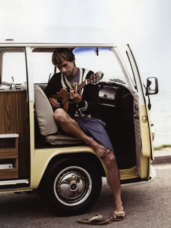 Barefoot man sits in the doorway of a vintage camper van playing guitar, with his sandals lying nearby on the asphalt road.