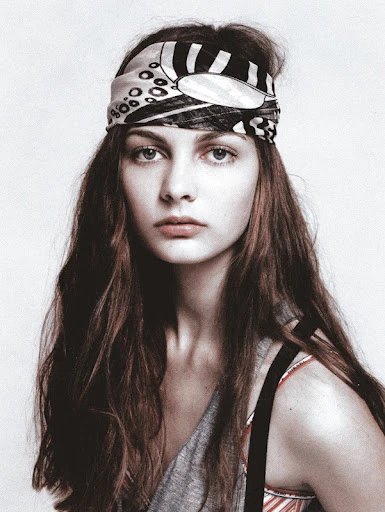 Close-up portrait of a fair-skinned young woman with long brown hair wearing a patterned headscarf looking directly at the camera.