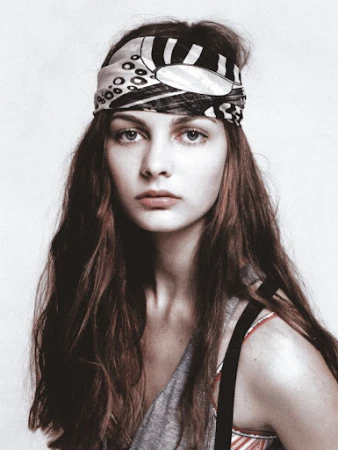 Close-up portrait of a fair-skinned young woman with long brown hair wearing a patterned headscarf looking directly at the camera.