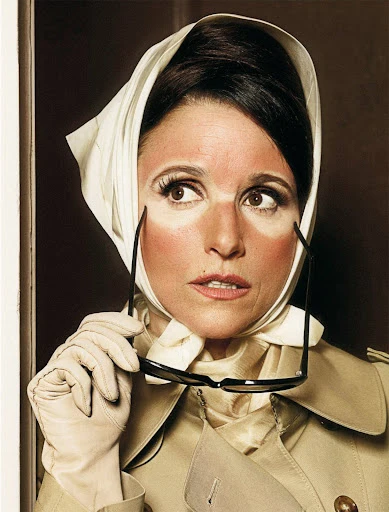 Woman with sunburn and headscarf holds sunglasses, wearing gloves and a trench coat in an indoor studio environment.