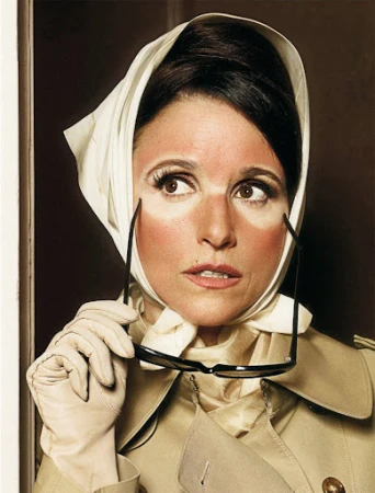 Woman with sunburn and headscarf holds sunglasses, wearing gloves and a trench coat in an indoor studio environment.