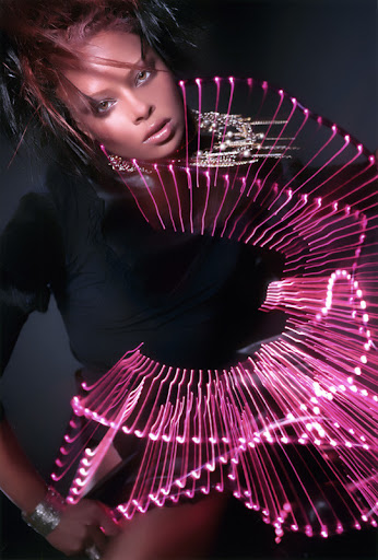 Portrait of a woman with dark hair and jewelry, wearing an illuminated collar. The background is dark and neutral in color.
