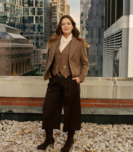 Drew Barrymore poses in a brown tweed vest and jacket with culottes and heels on a rooftop in an urban setting with buildings in the background.