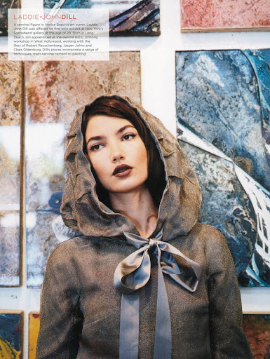 Portrait of a brunette fashion model wearing a hooded coat with a ribbon in front of abstract artwork in an indoor setting.