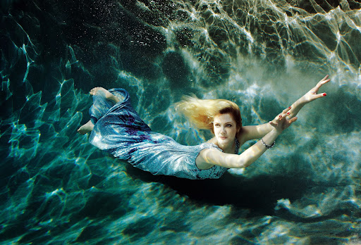 Woman in flowing dress underwater, floating with arms outstretched in pool with sunlight patterns on the turquoise water, blonde hair.