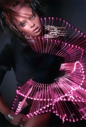 Portrait of a woman with styled dark hair and metallic jewelry, framed by bright pink light painting streaks in a dark studio setting.