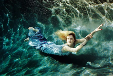 Woman in a sparkling blue dress swimming underwater, hair fanned out with rippled light patterns above