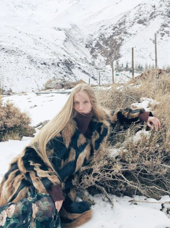 Long-haired blonde woman wearing a patterned faux fur coat in a snowy mountain landscape