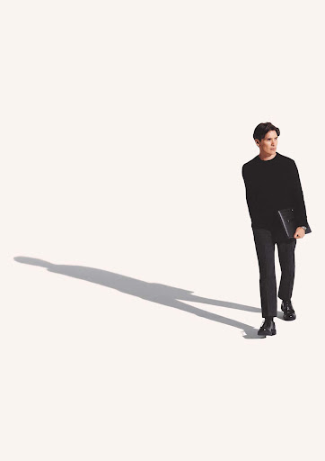A young adult male in a dark outfit walks and carries a laptop against a plain white background with his shadow.