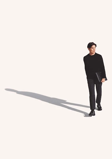 A young adult male in a dark outfit walks and carries a laptop against a plain white background with his shadow.