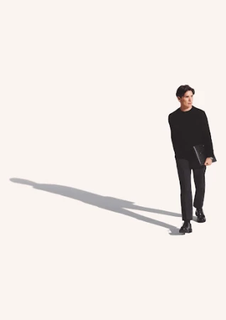 A young adult male in a dark outfit walks and carries a laptop against a plain white background with his shadow.