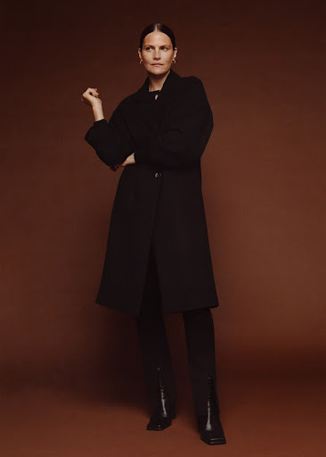 A woman in a black coat, black pants, and black heeled boots poses in front of a brown backdrop with one hand raised elegantly.