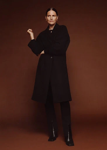 A woman in a black coat, black pants, and black heeled boots poses in front of a brown backdrop with one hand raised elegantly.