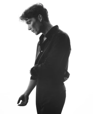 Black and white portrait of actor Cillian Murphy in a dark shirt against a bright seamless background.