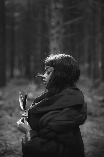 A young woman in a coat stands in a dark forest, holding a feather. Moody black and white portrait in a natural outdoor setting.