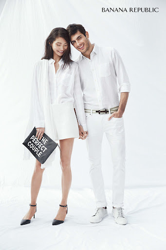 A casually dressed couple posing in a photo studio, the woman holding a clutch with text, set against a bright white backdrop.