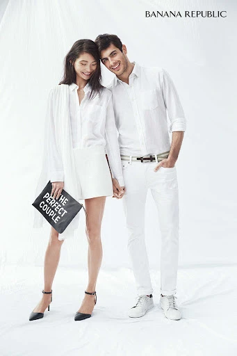 A casually dressed couple posing in a photo studio, the woman holding a clutch with text, set against a bright white backdrop.
