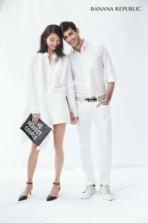 A casually dressed couple posing in a photo studio, the woman holding a clutch with text, set against a bright white backdrop.