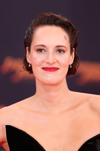 Close-up studio shot of Phoebe Waller-Bridge with short dark hair, wearing a black strapless dress and red lipstick against a dark background.