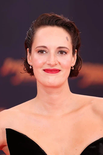 Close-up studio shot of Phoebe Waller-Bridge with short dark hair, wearing a black strapless dress and red lipstick against a dark background.