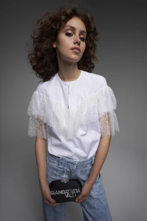 A woman with voluminous curly brown hair wears a white lace-trimmed top and holds a black clutch bag against a grey background.
