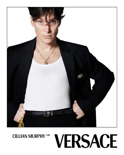 Cillian Murphy models Versace in a close-up studio shot. He wears a black blazer, white tank top, and gold necklace against a white background.